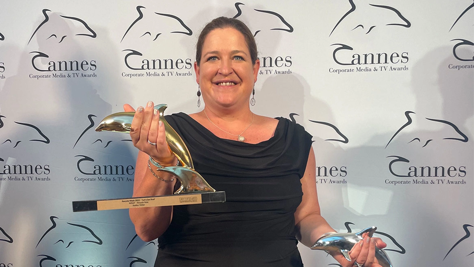 Radley Yeldar Head of film and animation Emily Clements smiling, wearing a black dress and holding a gold dolphin trophy in her right hand and a silver dolphin trophy in her left, standing in front of a step and repeat with Cannes Corporate Media & TV Awards branding