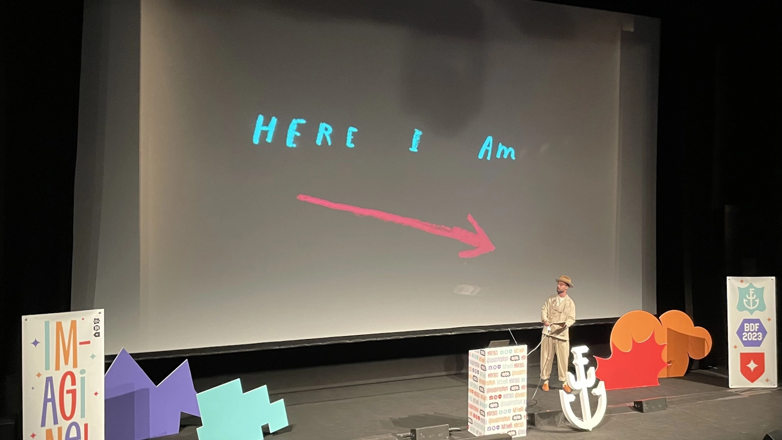 The stage at Birmingham Design Festival with the screen reading 'Here I Am' above an arrow that points to the speaker