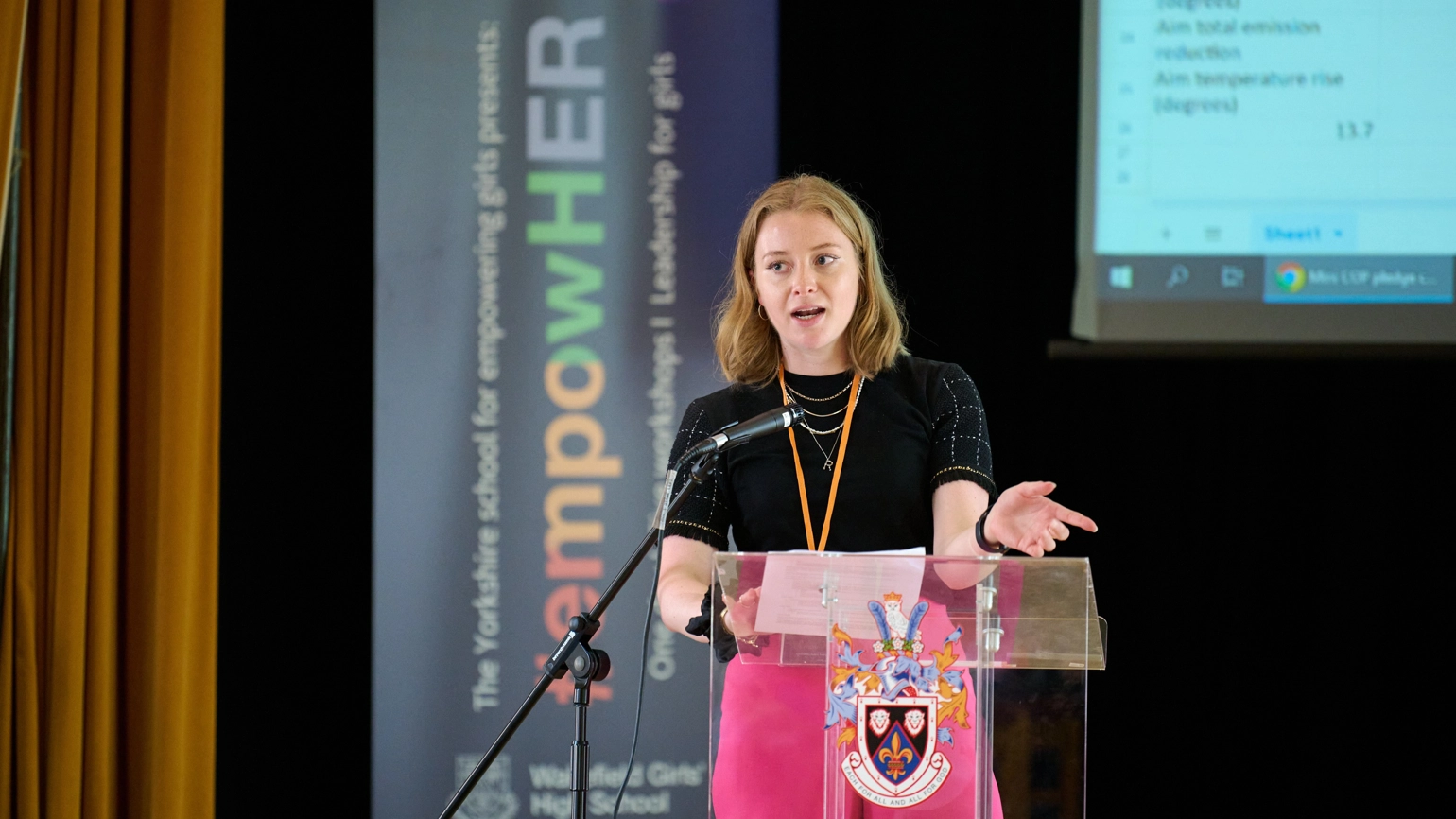 Rebecca Ward speaking in front of a podium at EmpowerHer event (branding in the background)
