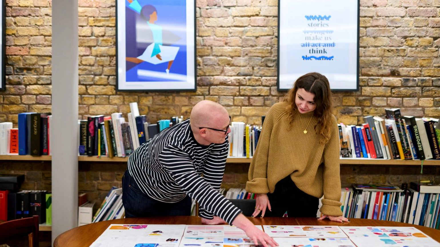 Two RY team members standing behind a desk, looking down large sheets of paper, the person on the left pointing towards the top. In the background are shelves of books and two large creative posters.