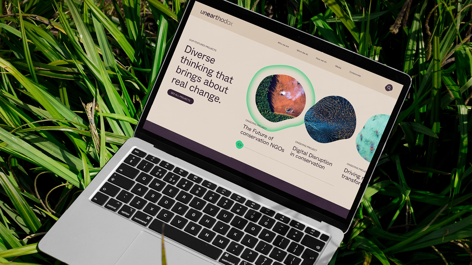 A computer sitting on a grass background, the screen shows Unearthodox's website, the title reading 'Diverse thinking that brings about real change'
