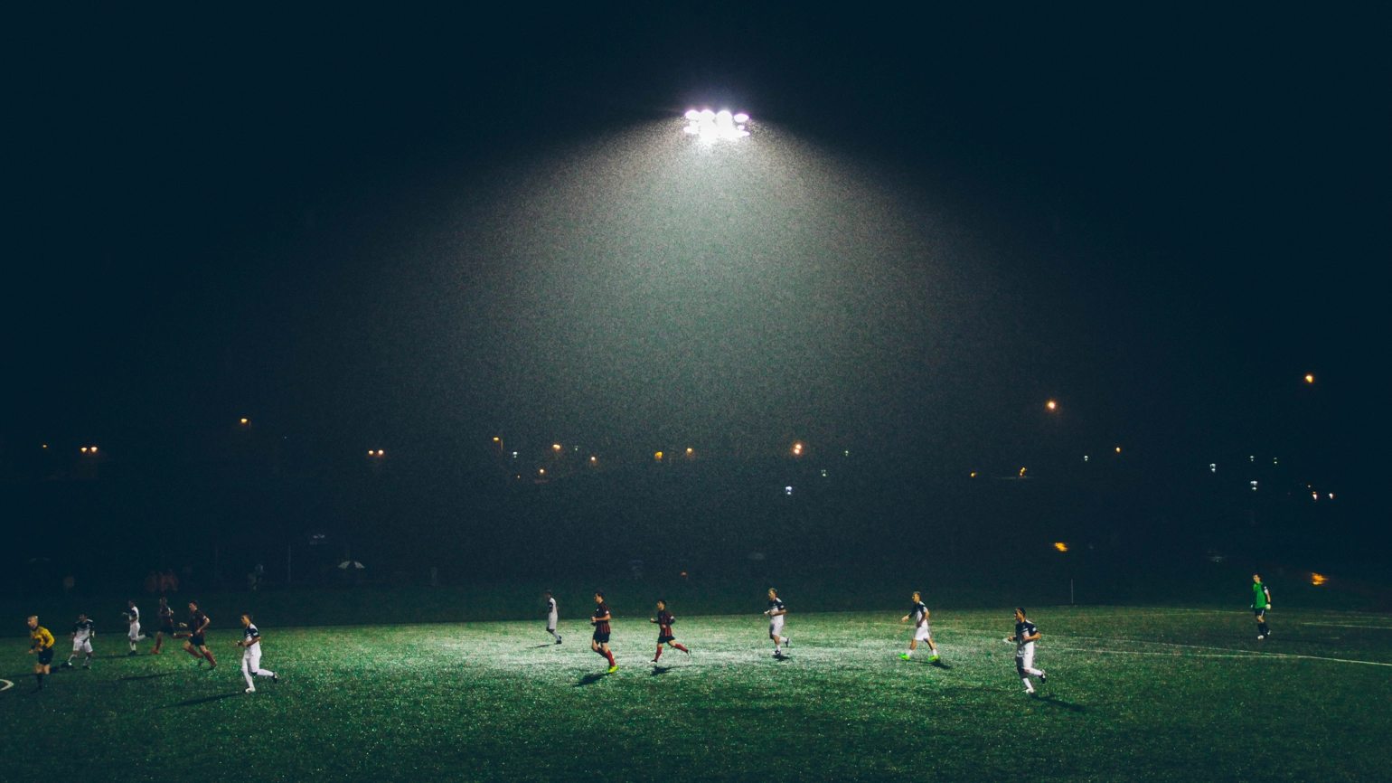 Players on a dark football pitch, lit by floodlights in the centre